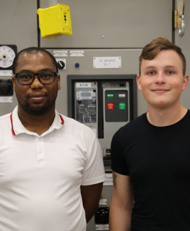 Nathanael stands on the left wearing a white shirt. Cody stands next to him wearing a black shirt. A control panel is in the background.