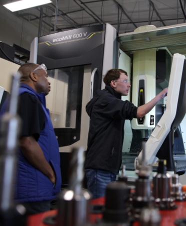 Tools sit in the foreground while a student wearing safety glasses and a black hoodie and jeans works on a machine. An instructor wearing a blue shirt watches.