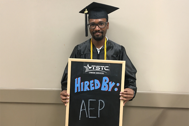 TSTC graduate posing in cap & gown holding a chalkboard showcasing their new employer.