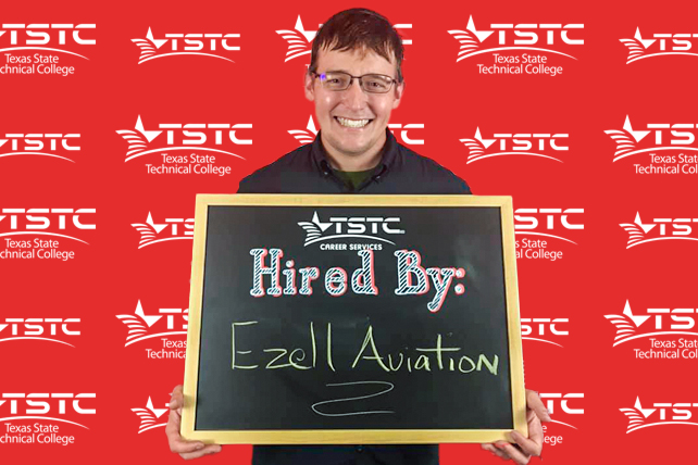 TSTC graduate posing in cap & gown holding a chalkboard showcasing their new employer.