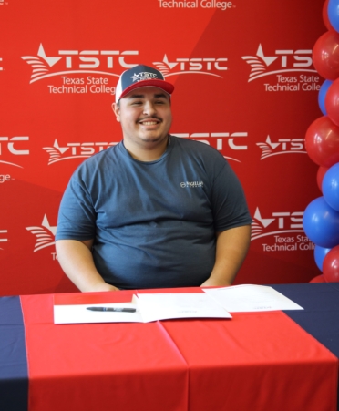Eddie sits at a table decorated in red and blue after signing papers. A TSTC banner rests in the background.