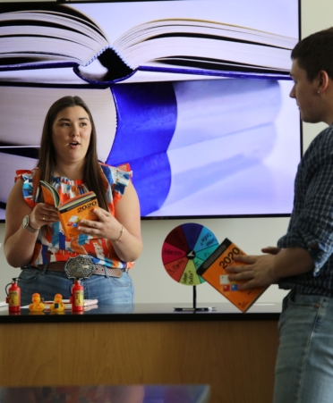 Kate wearing a colorful shirt holds open a book and talks with another student wearing a blue shirt.