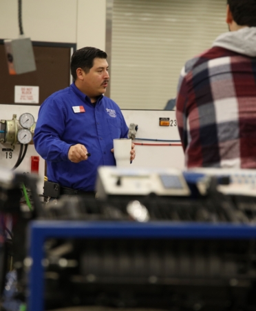 A man wearing a blue shirt speaks to a group of students with their backs turned from the camera. Equipment takes up the foreground.