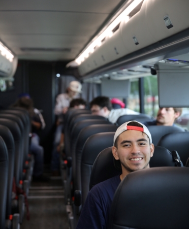 A student wearing a white ball cap backwards sits on a bus seat while students mill around in the background.