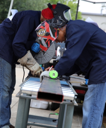 Two high school students wearing jeans, a blue shirt and protective gear work on a metal bar.