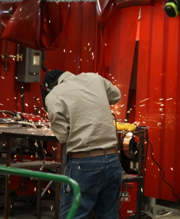 A student wearing jeans and a tan shirt welds something with his back turned. Sparks fly across the room.