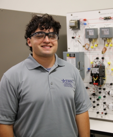 Dario stands in a gray TSTC polo with HVAC training equipment behind him.
