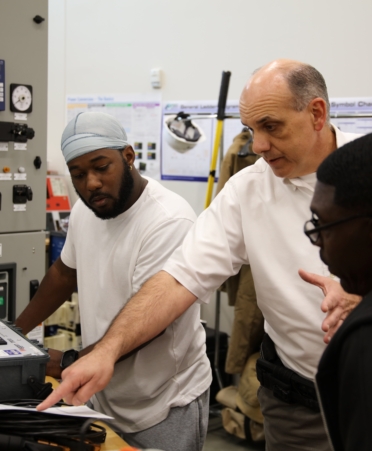 Two students and an instructor look at a some equipment and paperwork on a table. The instructor points at something on the paper.