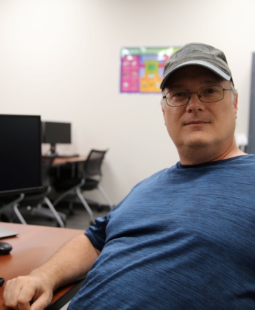Paul sits at a computer desk wearing a gray ballcap and a blue shirt.