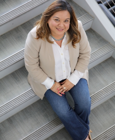 Larissa in a dress shirt and blazer, jeans, and dress shoes, sits on steps and looks at camera above her.