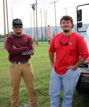 Mike stands, arms crossed, wearing a red shirt and a hat. Thomas stands next to him, hands in pockets, wearing a brighter red shirt.