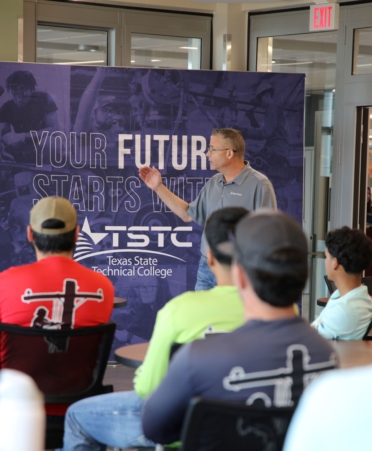 Rodney, wearing a grey polo, stands in front of a group of students mostly wearing TSTC lineworker shirts.