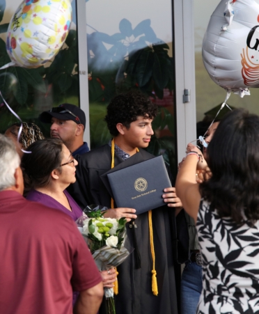 A Graduate wearing a gown and holding his diploma is surrounded by family and friends.